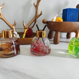 May include: Three glass bottles with gold lids and different coloured dried flowers inside. The bottles are sitting on a white surface.