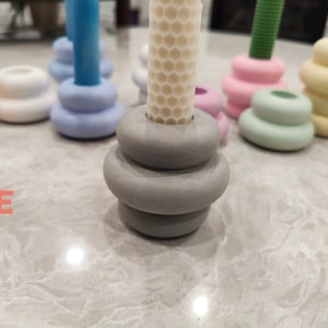 May include: A set of six candle holders in various pastel colors, including light blue, light green, light pink, and yellow. The candle holders are made of ceramic and have a stacked, rounded design. The candle holders are arranged on a white surface.