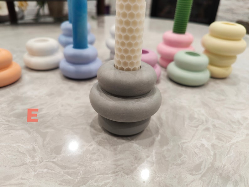 May include: A set of six candle holders in various pastel colors, including light blue, light green, light pink, and yellow. The candle holders are made of ceramic and have a stacked, rounded design. The candle holders are arranged on a white surface.