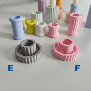 May include: Two concrete candle holders, one gray and one pink, with a fluted design. The gray holder has a smaller, inner cylinder. The pink holder is a single cylinder.