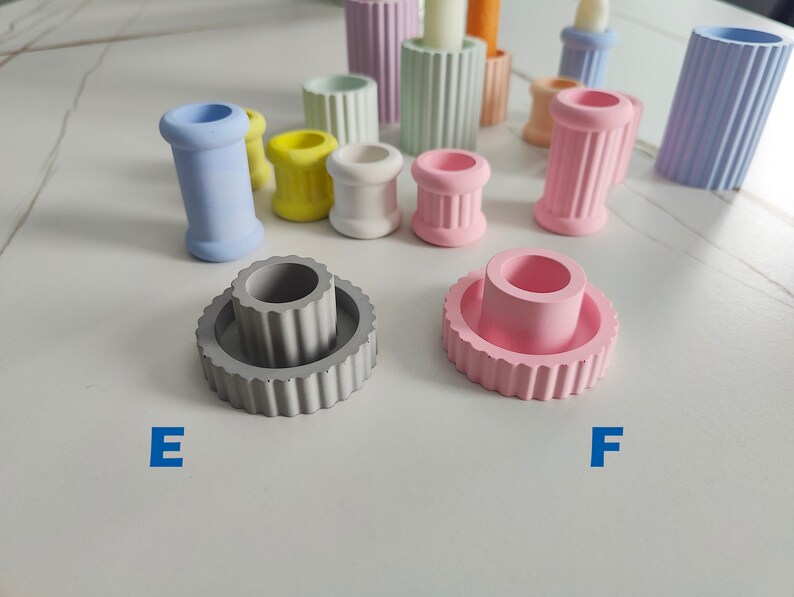 May include: Two concrete candle holders, one gray and one pink, with a fluted design. The gray holder has a smaller, inner cylinder. The pink holder is a single cylinder.