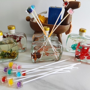 Puede incluir: Cuatro botellas de vidrio con flores secas de colores y líquido transparente, utilizadas para difundir aceites esenciales. Las botellas están dispuestas sobre una superficie blanca con palitos blancos y cañas difusoras cuadradas de colores.