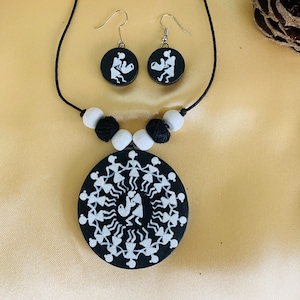 May include: A black necklace and earring set featuring a Warli art design. The necklace has a large circular pendant with a white tribal design and white and black beads. The earrings are small, round, and feature a similar design.