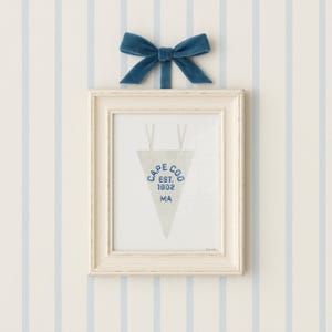 May include: A framed pennant with the text "CAPE COD EST. 1602 MA" in blue, hanging below a blue velvet bow. The frame is off-white, and the background is a striped pattern of light blue and white.