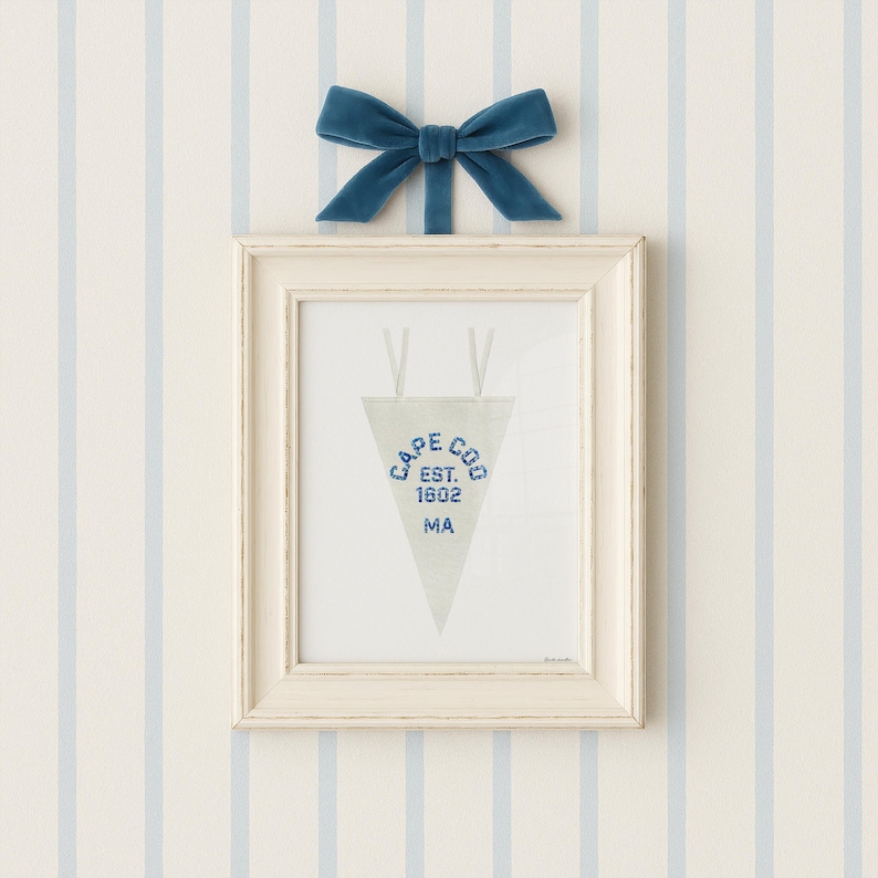May include: A framed pennant with the text "CAPE COD EST. 1602 MA" in blue, hanging below a blue velvet bow. The frame is off-white, and the background is a striped pattern of light blue and white.