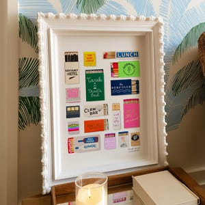 May include: A white framed artwork featuring a collection of colorful matchboxes. The matchboxes have various designs and text, including "Montauk Hotel," "Lunch," and "Clam Bar." The frame has a decorative, textured border.