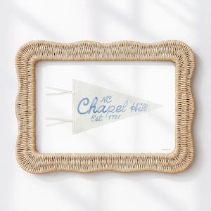 Puede incluir: Una impresión enmarcada con un banderín azul claro con las palabras "Chapel Hill" y "NC Est. 1758" en una fuente de escritura. El banderín está sobre un fondo blanco y enmarcado en un marco rectangular tejido de color marrón claro.