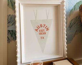 Puede incluir: Impresión enmarcada en blanco con un banderín con la inscripción "AUSTIN EST. 1839 TX" en letras rojas. El marco tiene un borde decorativo. El fondo tiene un diseño de palmeras.