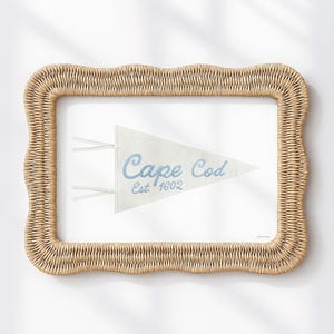 Puede incluir: Una obra de arte mural rectangular con un banderín de color claro con las palabras "Cape Cod" en escritura azul y "Est. 1602". El banderín está sobre un fondo blanco y enmarcado por un marco tejido, marrón claro, con bordes ondulados.