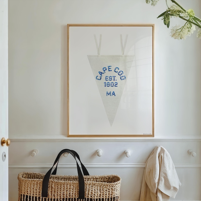 May include: A framed print featuring a white pennant with the text "CAPE COD EST. 1602 MA" in blue. The print is in a gold frame and hangs on a white wall. A woven basket and coat are below.