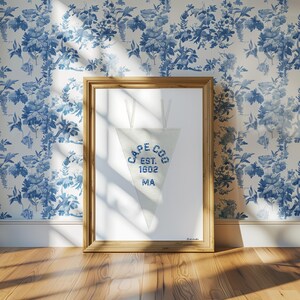 May include: A framed pennant with the text "CAPE COD EST. 1602 MA" in blue lettering. The pennant is white and triangular, hanging from a wooden frame. The background features a blue and white floral wallpaper.