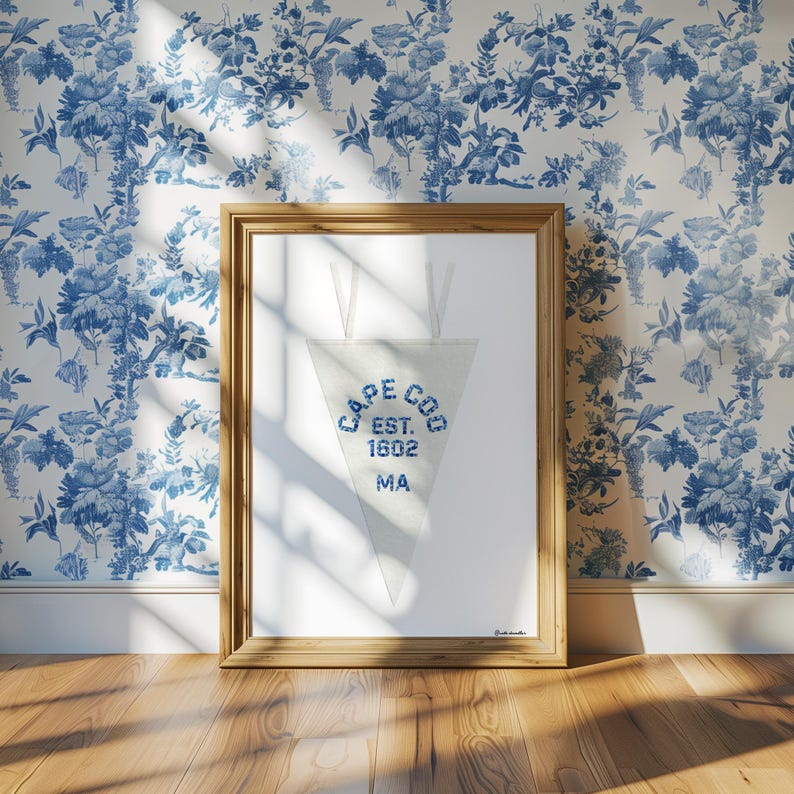 May include: A framed pennant with the text "CAPE COD EST. 1602 MA" in blue lettering. The pennant is white and triangular, hanging from a wooden frame. The background features a blue and white floral wallpaper.