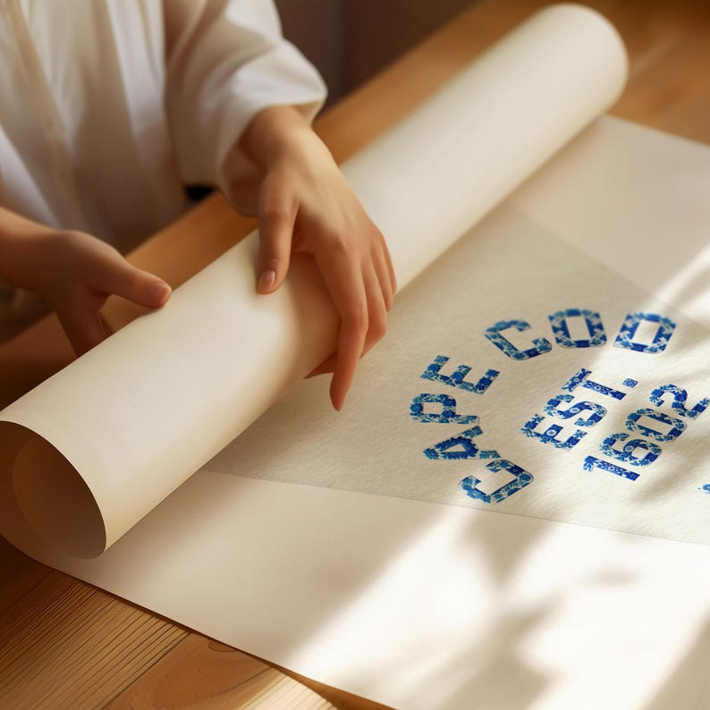 May include: A rolled-up white paper print with blue text that reads "CAPE COD EST. 1602". The paper is being unrolled on a wooden surface. The text is in a decorative font.