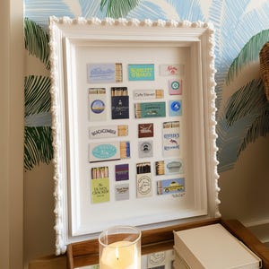 May include: A white framed display featuring a collection of vintage matchboxes. The matchboxes are arranged inside the frame, showcasing various designs and logos. The frame has a decorative edge and is displayed on a wooden surface.