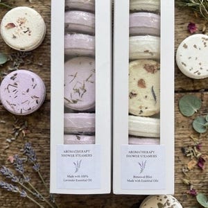 May include: Two vertical white rectangular boxes containing stacked round shower steamers. The left box label reads AROMATHERAPY SHOWER STEAMERS Made with 100% Lavender Essential Oil. The right box label reads AROMATHERAPY SHOWER STEAMERS Botanical Bliss Made with Essential Oils.