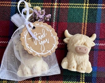 May include: A cream-colored Highland cow-shaped soap figurine next to a white organza drawstring bag containing another identical soap, topped with dried purple and yellow flowers and a round brown paper tag with the text Thank you.
