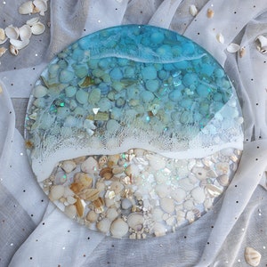 May include: A round, clear resin tray with a blue and white wave design. The tray is filled with small seashells and glitter.