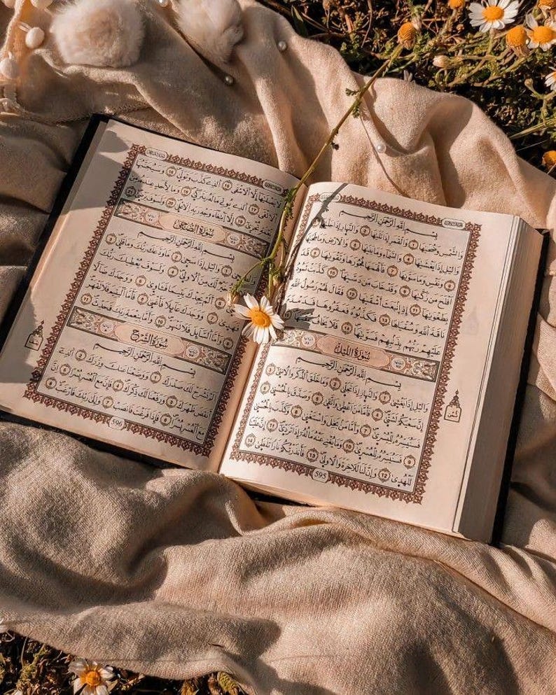 May include: An open Quran, a holy book of Islam, lies on a beige fabric with a white flower on the page. The fabric is on a bed of green grass with white flowers.