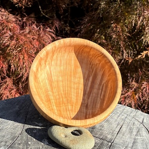 Peut inclure: Un bol en bois clair avec un motif de grain de bois visible. Le bol est posé sur une surface en bois vieilli. Une petite pierre grise avec un trou est devant le bol. L'arrière-plan présente un feuillage rouge et vert.