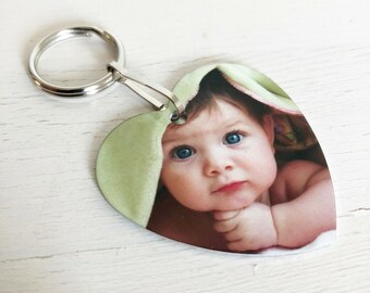 May include: A heart-shaped metal keychain with a photo of a baby girl with blue eyes. The baby is wearing a green blanket and is looking at the camera.