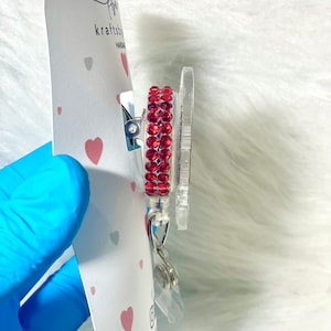 May include: A clear badge reel with a red rhinestone border. The reel is attached to a white card with a heart pattern and the text "kraftsby". The badge reel has a silver clip and a clear plastic retractable cord.