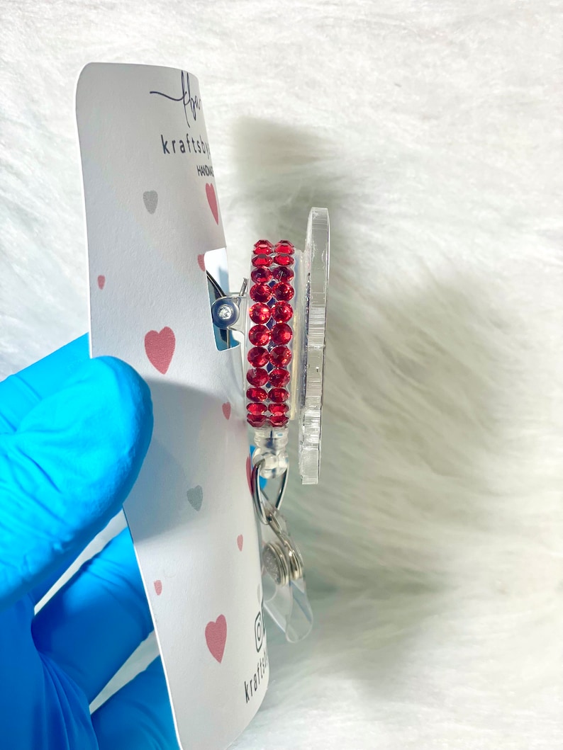 May include: A clear badge reel with a red rhinestone border. The reel is attached to a white card with a heart pattern and the text "kraftsby". The badge reel has a silver clip and a clear plastic retractable cord.