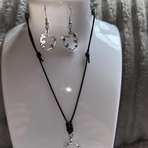 Op de afbeelding: Een zilverkleurige sieradenset met een ketting en oorbellen. De ketting heeft een zwart koord met een zilveren bladhanger. De oorbellen zijn bijpassende zilveren bladvormen. De set wordt gepresenteerd op een witte mannequinhoofdstuk.