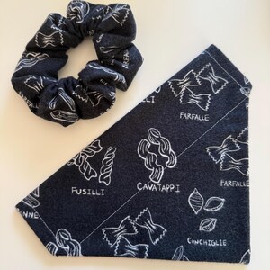 May include: A dark blue scrunchie and a matching bandana featuring a white pasta design. The bandana is triangular and displays various pasta shapes with their names, including fusilli and farfalle.