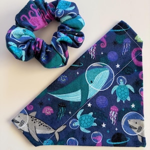 May include: A navy blue scrunchie and a triangular pet bandana. Both items feature a space-themed design with whales, turtles, and sharks in astronaut helmets, along with planets and jellyfish. The scrunchie has teal, purple, and pink accents.