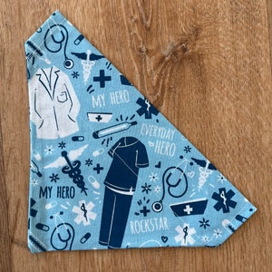 May include: Blue bandana with a white and blue pattern of medical symbols, including a stethoscope, a thermometer, a bandage, a pill, a cross, a caduceus, and a doctor's coat. The bandana also has the text "My Hero", "Everyday Hero", and "Rockstar".