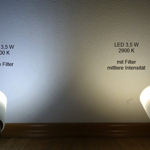 Peut inclure: Deux ampoules LED blanches avec des abat-jours en verre dépoli, l'une avec un filtre et l'autre sans. Le texte sur le mur indique "LED 3,5 W 2900 K" et "ohne Filter" pour la lumière sans filtre et "mit Filter mittlere Intensität" pour la lumière avec un filtre.
