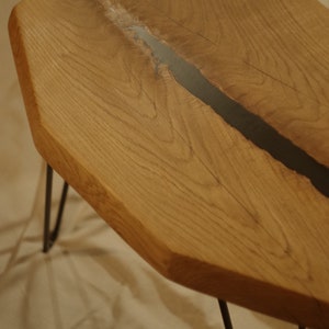 Op de afbeelding: Een houten tafel met een zwarte harsinleg. De tafel heeft een unieke, onregelmatige vorm en wordt ondersteund door drie zwarte haarspeldpoten.