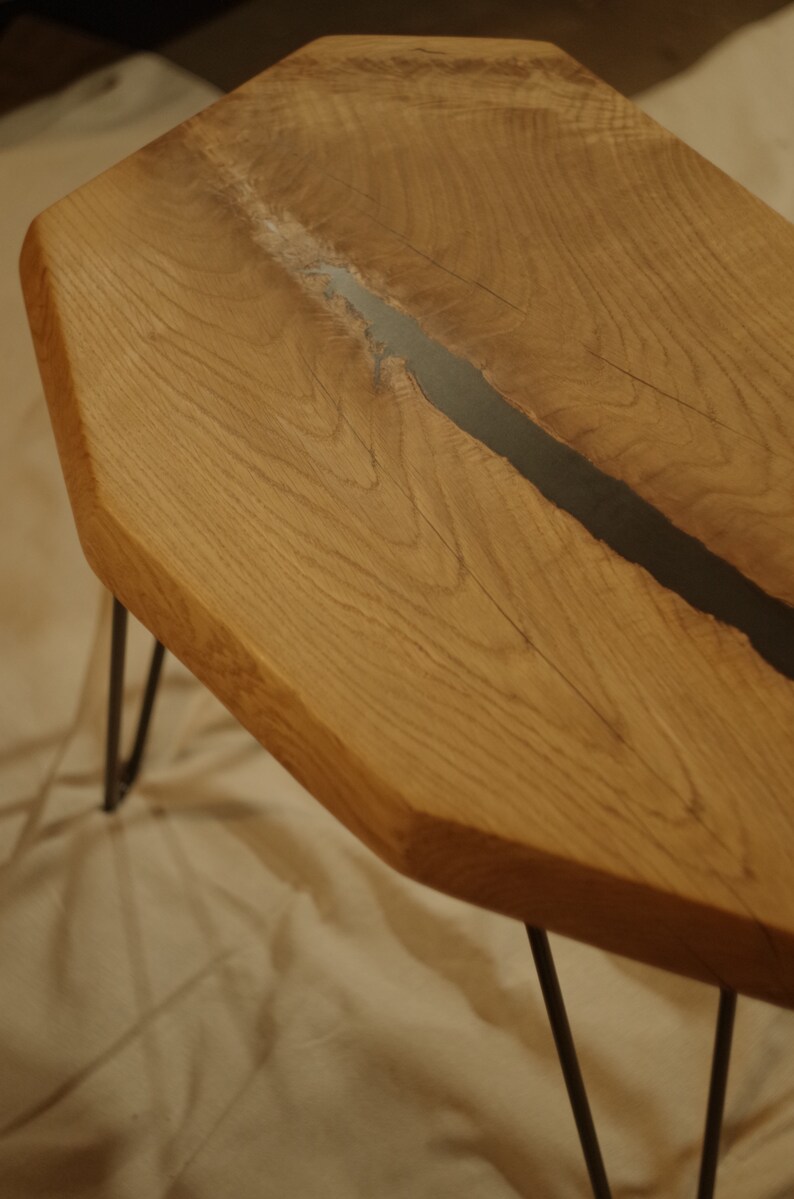 Op de afbeelding: Een houten tafel met een zwarte harsinleg. De tafel heeft een unieke, onregelmatige vorm en wordt ondersteund door drie zwarte haarspeldpoten.