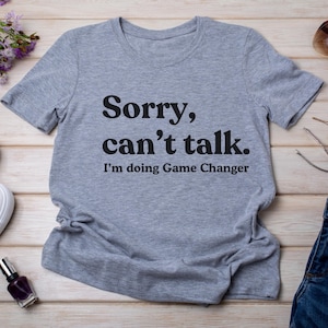 Könnte beinhalten: Hellgraues T-Shirt mit dem schwarzen Text "Sorry, can't talk. I'm doing Game Changer". Das Shirt liegt flach auf einer hellen Holzoberfläche, mit Accessoires wie Sonnenbrille und Nagellack.