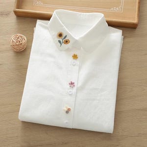 May include: White collared shirt with embroidered sunflowers on the collar and decorative flower-shaped buttons. The shirt is folded neatly on a wooden surface, showcasing a clean and delicate design. The buttons are yellow, pink, and a mix of colors.