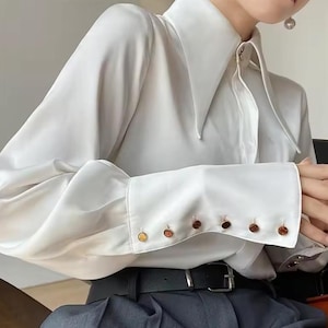 May include: White long-sleeve blouse with a large, pointed collar and button cuffs. The cuffs have multiple decorative buttons.