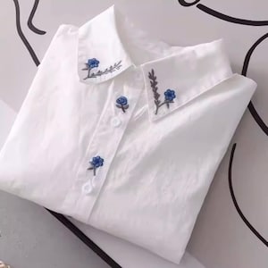 May include: A white button-down shirt with a pointed collar. The collar and button placket are adorned with delicate blue floral embroidery. The shirt is folded neatly, showcasing the details of the design.