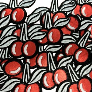 May include: A collection of embroidered patches featuring red cherries with white highlights and black outlines. Each cherry has a green stem and white and black striped leaves. The patches are densely packed together.