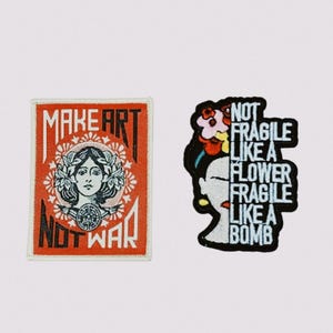 May include: Two embroidered patches. One is rectangular with the words "MAKE ART NOT WAR" and a woman's face. The other patch is a woman's face with flowers and the words "NOT FRAGILE LIKE A FLOWER FRAGILE LIKE A BOMB".