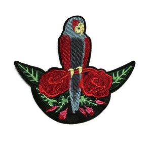 May include: Embroidered patch featuring a parrot with red and gray feathers perched above two red roses. The patch has a black crescent-shaped base with green leaves and stems. The parrot has a yellow beak.