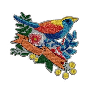 May include: Embroidered patch featuring a colorful bird perched on a branch with flowers and leaves. The bird has blue, yellow, and red feathers. An orange banner is woven through the floral arrangement. The patch has a gray border.