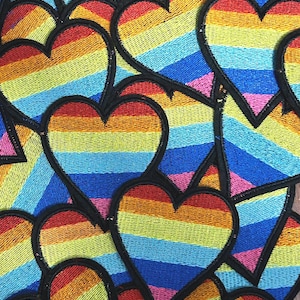 May include: A collection of embroidered heart patches in a rainbow color scheme. Each heart features horizontal stripes of red, orange, yellow, green, blue, and purple, outlined in black. The patches are densely packed together.