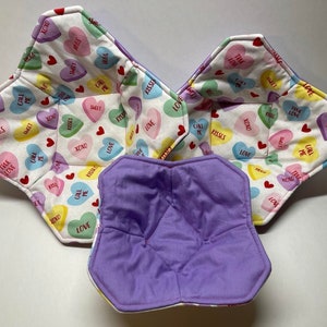 May include: Three fabric bowls with a white background and a pattern of colorful candy hearts with text such as "Call Me", "Sweet", "XOXO", "Kisses", and "True Love". The bowls are folded and have a purple lining.