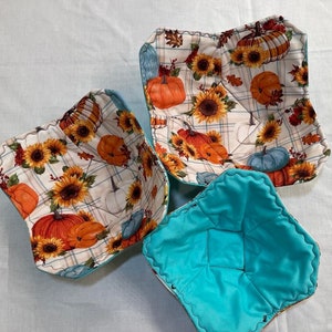 May include: Three fabric bowl cozies. Two are square and feature a white background with a plaid pattern, sunflowers, and pumpkins. The third is round and turquoise.