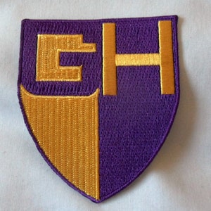 以下が含まれることがあります： 「EH」の文字が入った、盾型の刺繍ワッペン。ワッペンは垂直に分割され、左側は金色、右側は紫です。文字は金色で、背景は紫です。