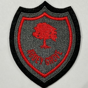 Peut inclure: Un écusson brodé gris avec un contour rouge en forme de bouclier. L'écusson représente un arbre rouge avec un tronc épais et une inscription rouge qui dit "Abbey Grove".