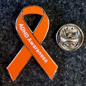 May include: Orange awareness ribbon pin with the words "ADHD Awareness" printed on it. The pin has a silver backing.