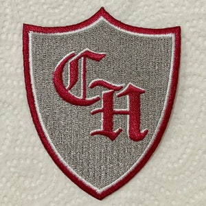 May include: Embroidered shield-shaped patch with a grey background and a red border. The patch features the letters "CH" in a decorative red font. The patch is likely for a school or organisation.