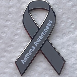 May include: A grey ribbon badge with the words "Asthma Awareness" in white lettering. The badge has a silver border and a glossy finish. It is a symbol of support and awareness.