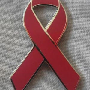 May include: A red awareness ribbon badge with a gold-coloured trim. The ribbon is a symbol of support and solidarity. The badge is made of metal and has a glossy finish. The ribbon is a classic design.